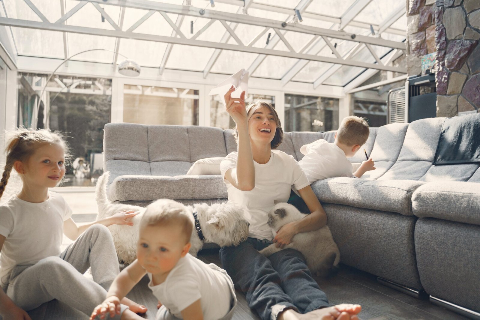 A family enjoys leisure time in a sunlit living room; a young woman and a child play with a paper airplane on a gray sofa, a toddler sits on the floor, and a second child draws in the background, accompanied by a white dog and a cat, suggesting a cozy, casual family moment at home.