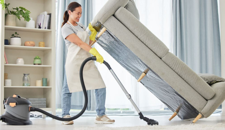 Professional cleaner vacuuming underneath an elevated sofa during a deep cleaning session in a bright living room