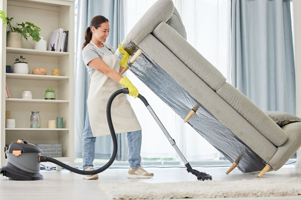 Professional cleaner vacuuming underneath an elevated sofa during a deep cleaning session in a bright living room