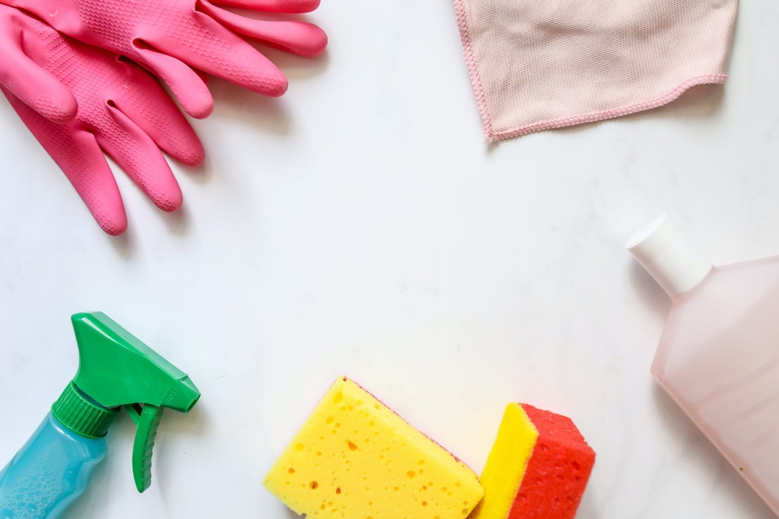 A flat lay of cleaning supplies on a white surface, including pink rubber gloves, a green spray bottle, a pink microfiber cloth, and two sponges, one yellow and one red.