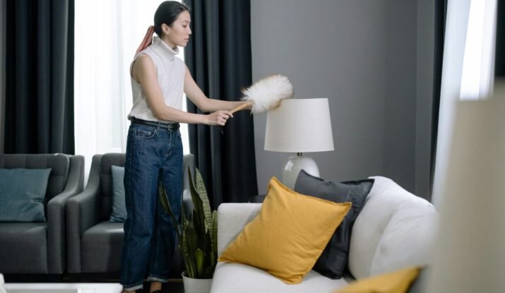 A person dusting a lampshade with a fluffy duster in a modern living room, exemplifying daily cleaning tasks.