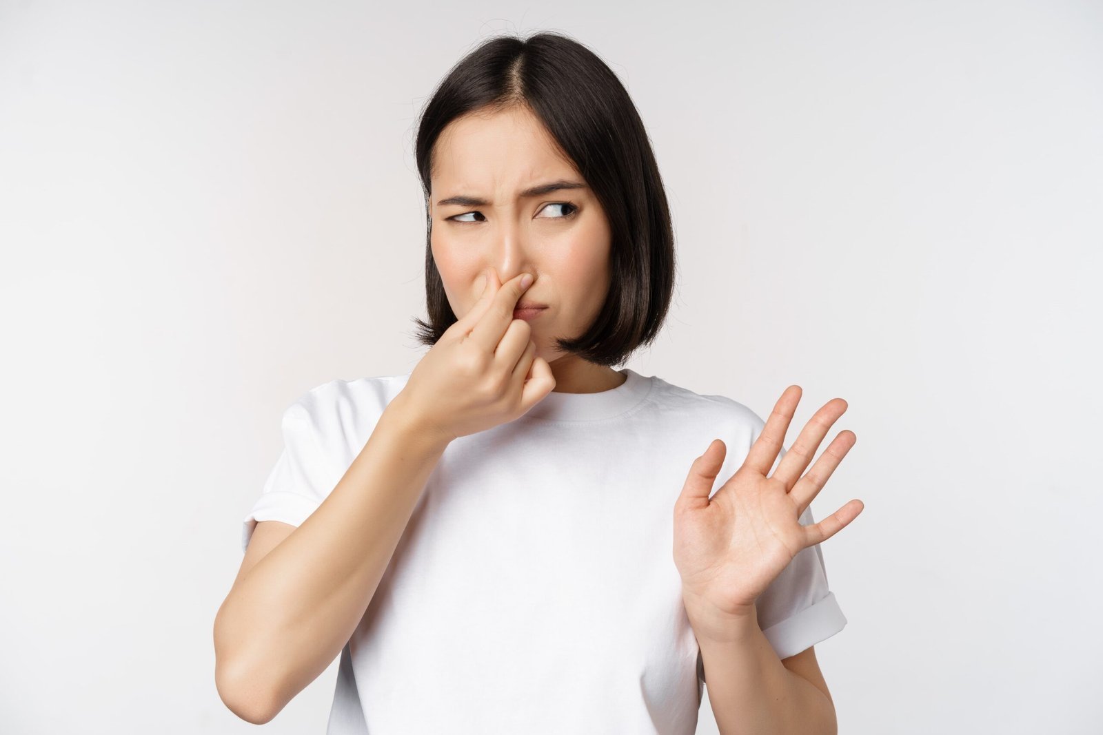 A woman in a white t-shirt is making a displeased face and holding her nose with one hand, indicating a bad smell, while gesturing stop with her other hand.