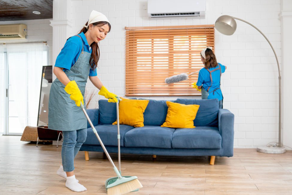 Two people in blue aprons and yellow gloves cleaning a living room, one sweeping the floor and the other dusting a window blind.