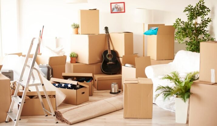 A room filled with unpacked moving boxes, various household items, and a rolled-up rug, indicating a moving day scene with belongings yet to be organized and set up in a new home