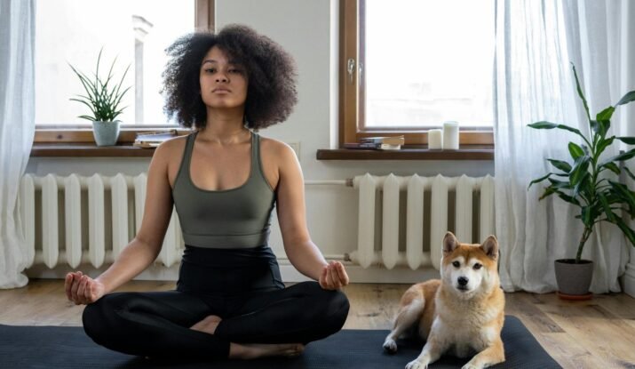A woman in a meditative pose practicing yoga on a mat in a well-lit room, accompanied by a Shiba Inu dog, with indoor plants contributing to a serene and healthy home environment.